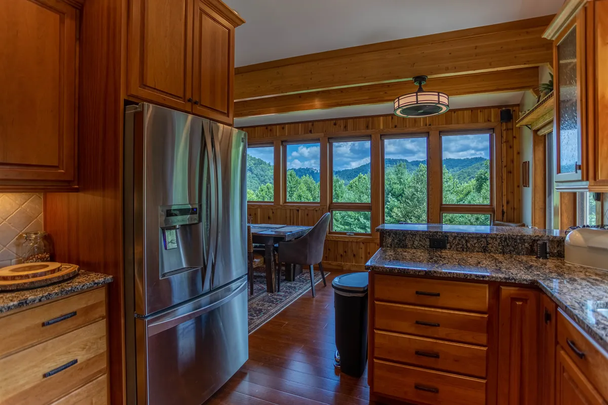 A spacious kitchen featuring stainless steel appliances, granite countertops, wood cabinetry, and a large window offering mountain vistas. A dining table is also visible, emphasizing a seamless flow between cooking and dining areas.