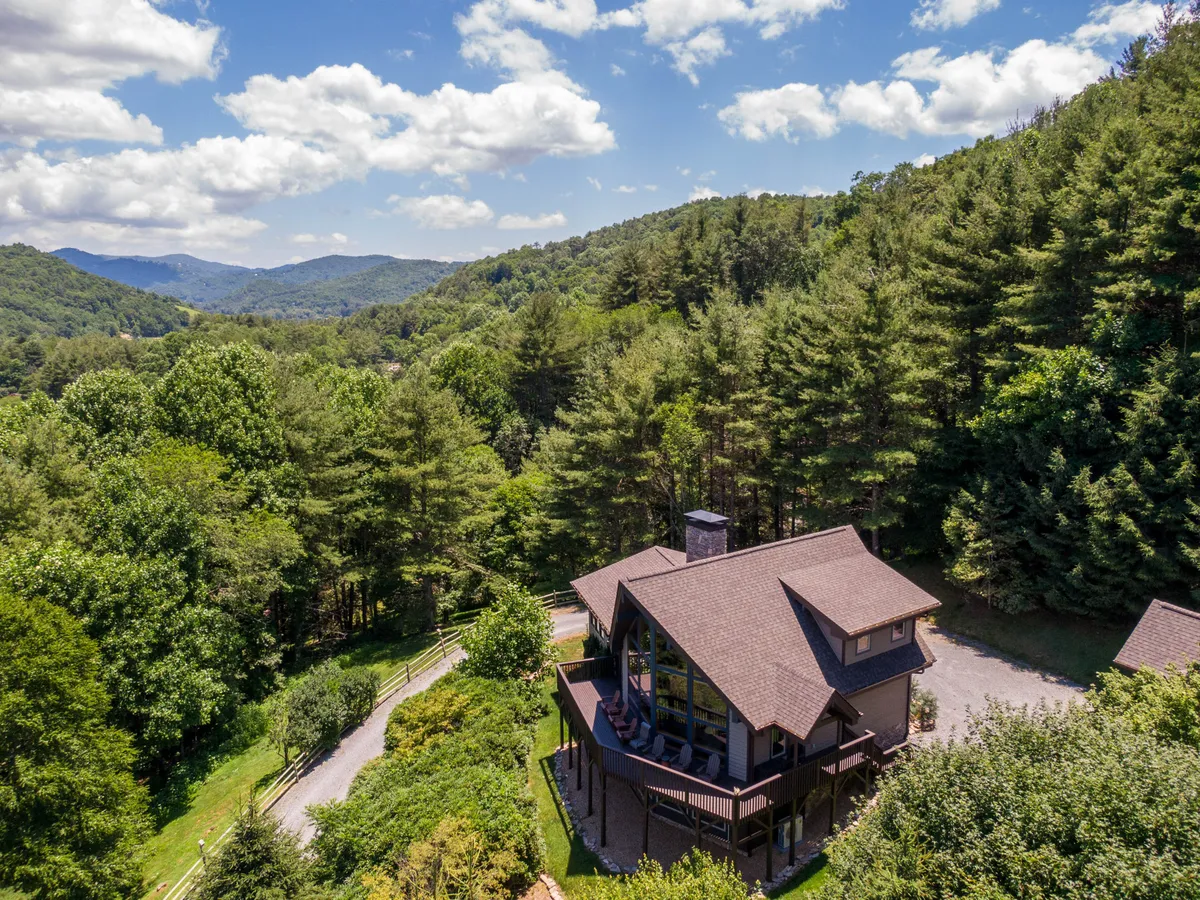 A wide exterior shot of the house enveloped by lush trees and offering expansive mountain views. The deck is visible, ideal for relaxing or enjoying the scenery after a day of adventure. The mood is peaceful, capturing the natural beauty and proximity to outdoor activities.