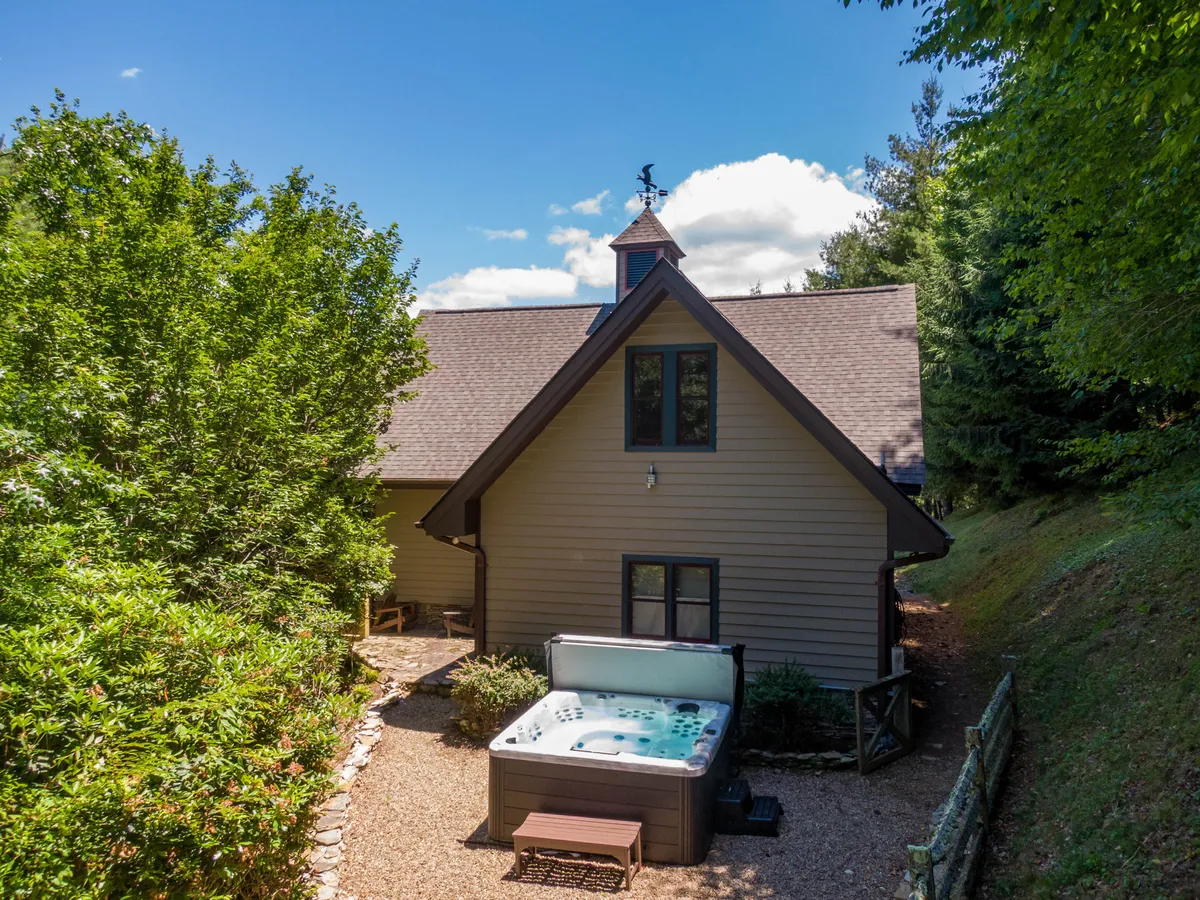 This image prominently features the hot tub set against lush greenery with the house facade in the background. The scene suggests a tranquil and private spot for relaxation, with the hot tub large enough for multiple people to enjoy. The context of the surrounding foliage conveys a sense of seclusion and natural beauty, ideal for unwinding after outdoor activities.