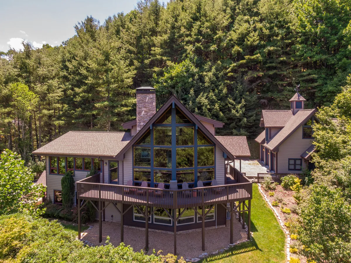 The image showcases the inviting exterior of a spacious cabin nestled in the lush greenery of Sugar Grove, NC. Large glass windows reflect the surrounding forest, while a wide wooden deck offers plenty of space for outdoor dining and relaxation. The prominent stone chimney hints at cozy fires inside, and the serene setting is perfect for families or groups to unwind and enjoy nature.