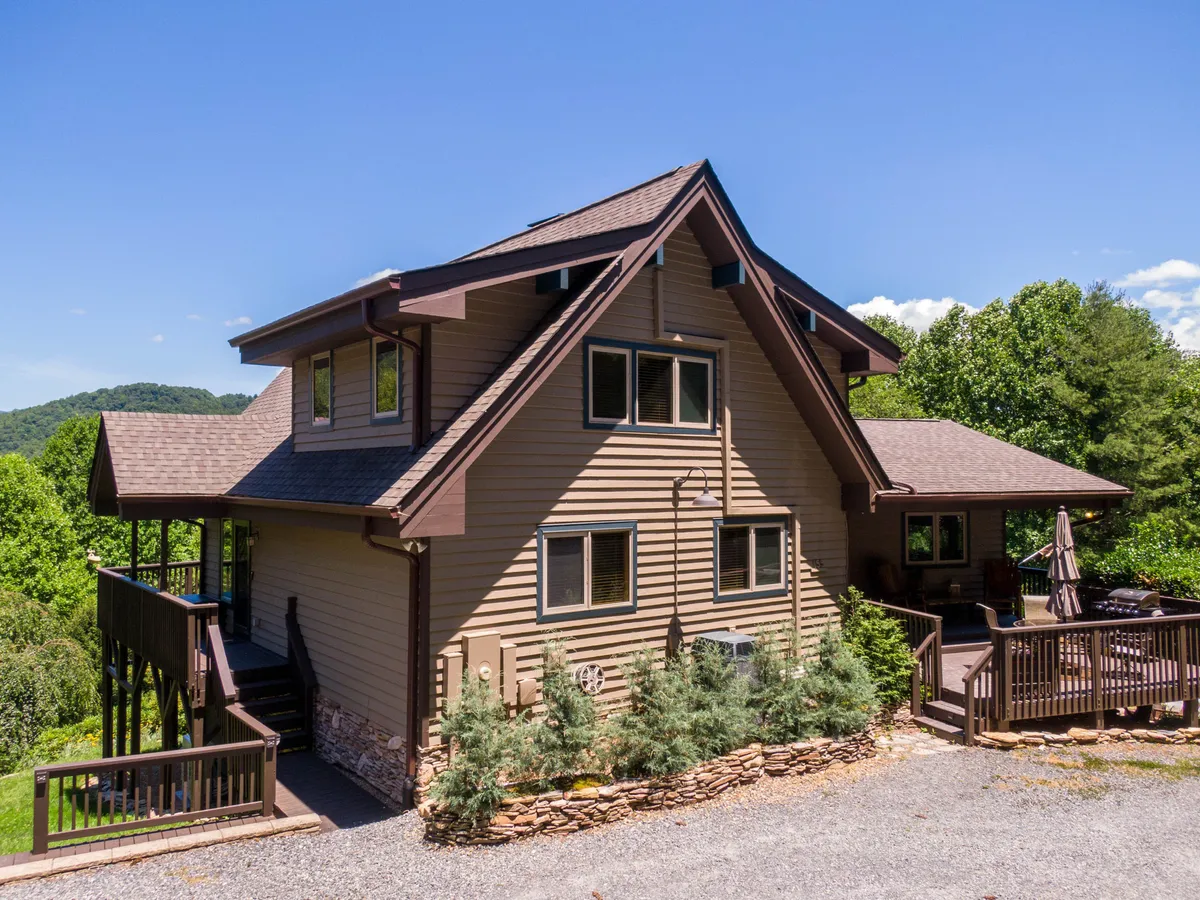 The exterior of the mountain outpost, featuring the house's facade, a spacious deck area, and inviting landscaping. The setting is serene and surrounded by nature, hinting at relaxing outdoor gatherings and connection to the landscape.