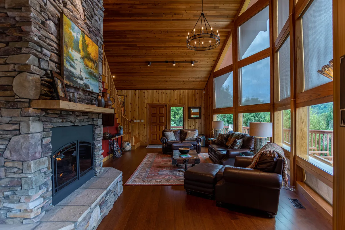 A warm, inviting living room with a prominent stacked stone fireplace at its center. Large floor-to-ceiling windows flood the space with natural light and frame unobstructed views of rolling mountains and sky. Comfortable seating is arranged to encourage conversation and relaxation, while a wooden ceiling adds rustic charm.