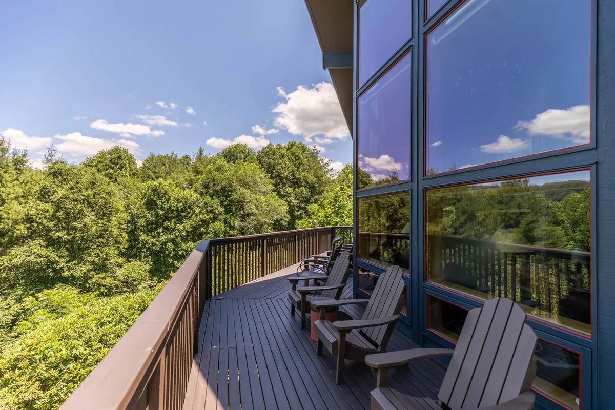 A spacious wooden deck furnished with Adirondack chairs overlooks a lush, tree-filled yard. Large windows from the house provide a seamless connection to the outdoors, making this a perfect spot to relax and watch over pets playing in the fenced yard below.