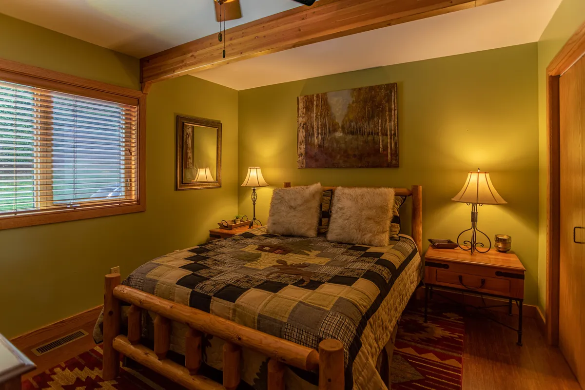 A warm, inviting bedroom with a rustic log bed, decorative pillows, nightstands with lamps, and framed artwork. The room is bathed in gentle light, offering a cozy and comfortable atmosphere perfect for restful nights.