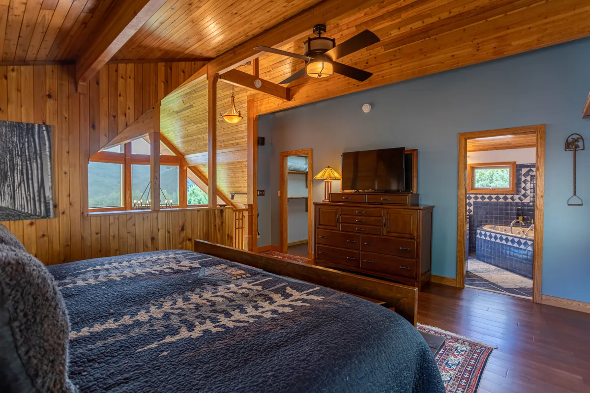 This image showcases a serene and comfortable bedroom with a bed featuring a patterned throw, large windows framing a stunning mountain view, wooden ceiling and walls, and a dresser with a television. The space exudes tranquility and is clearly designed for relaxation.